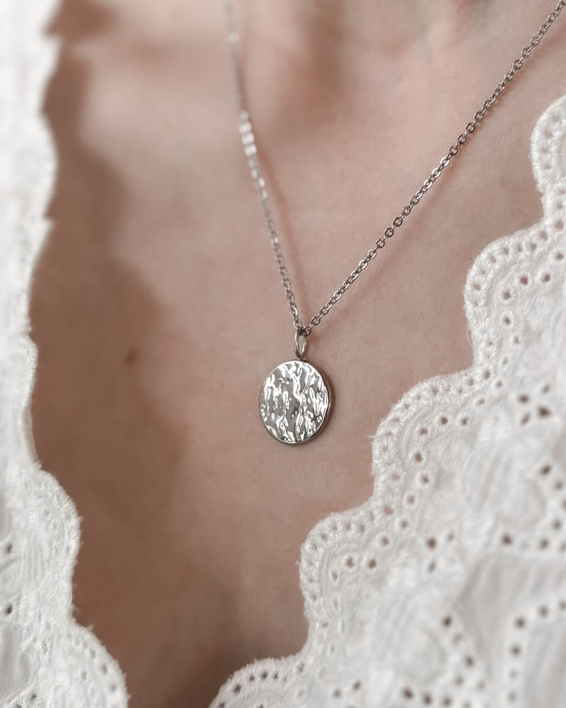 MOON QUOTE necklace in silver-coloured stainless steel with hammered round coin facing out, worn around the neck over a white lace top