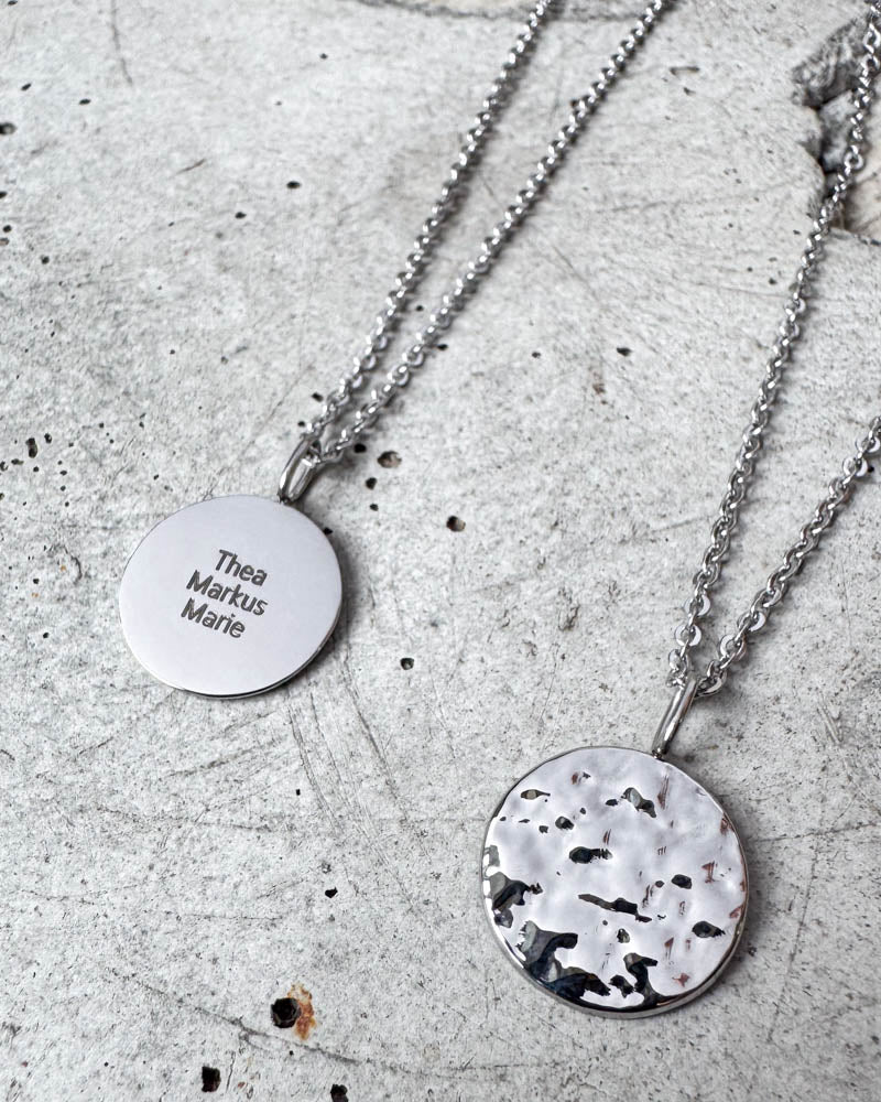MOON QUOTE name necklace in silver-coloured stainless steel engraved with names "Thea Markus Marie", hammered side alongside, on concrete
