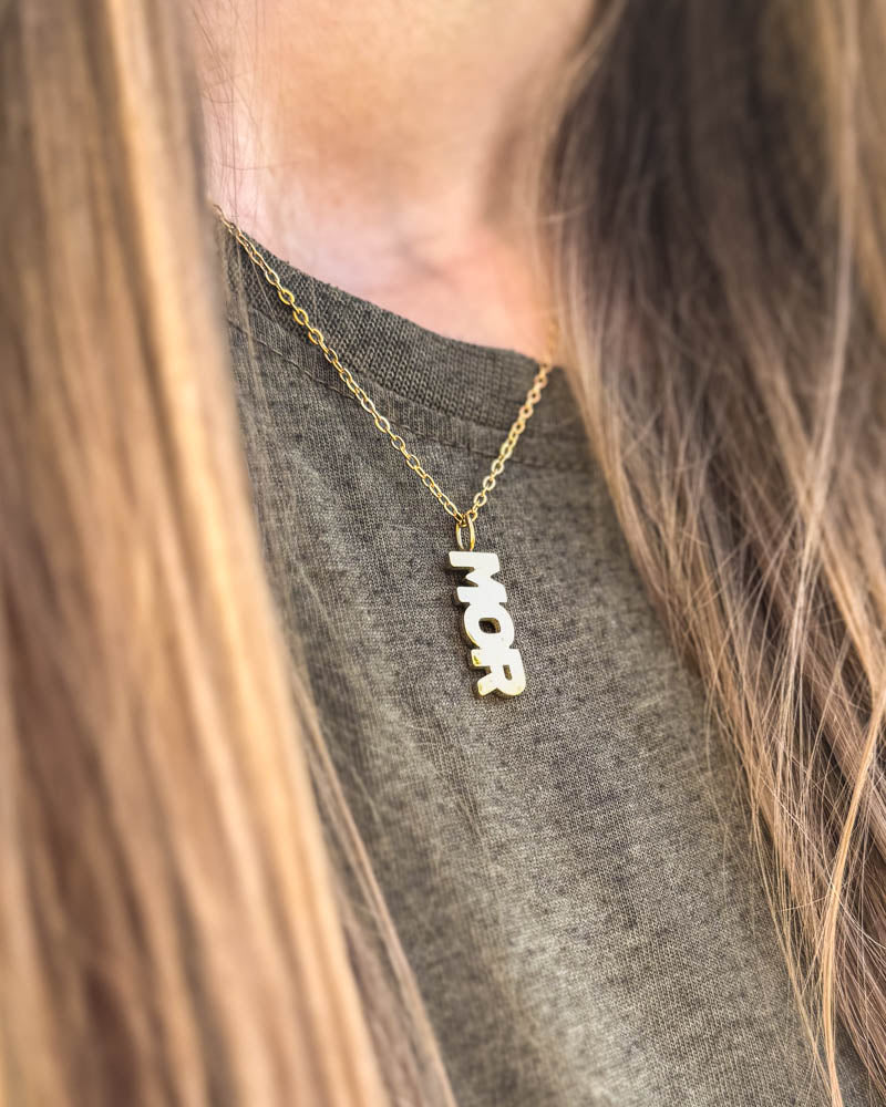 MOM necklace in gold-plated stainless steel with "MOR" letter pendant, worn around the neck over a grey t-shirt