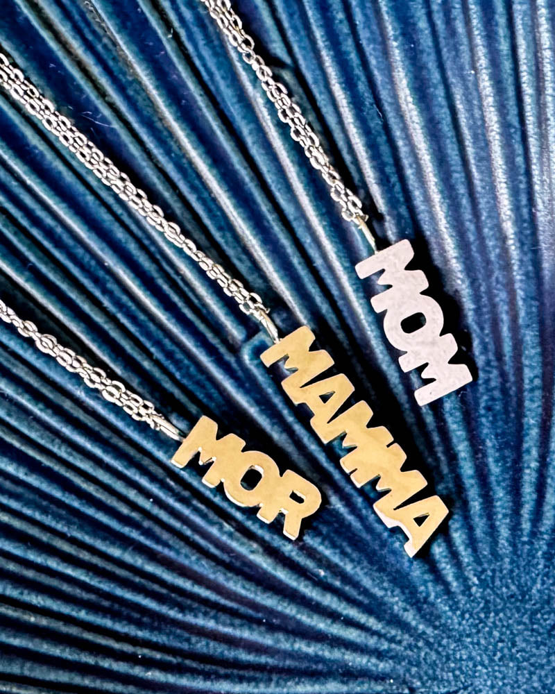 MOM necklaces with letter pendants "MOR" and "MAMMA" in gold-plated and "MOM" in silver-coloured stainless steel, on blue ribbed ceramic