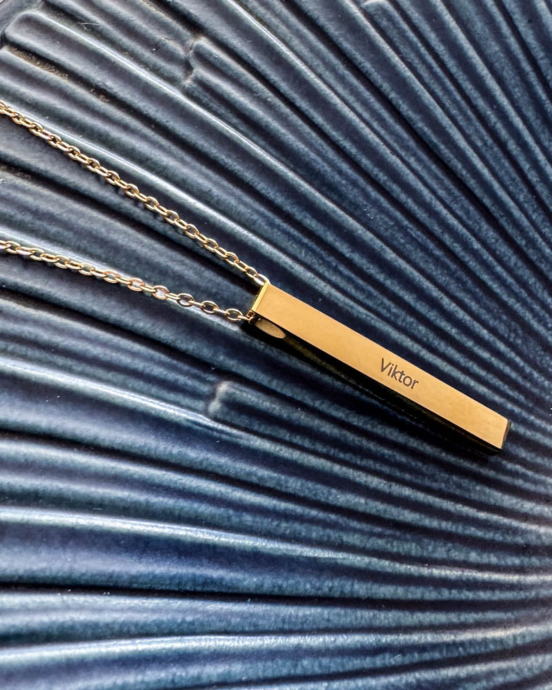 NEO name necklace in gold-plated stainless steel with the name "Viktor" engraved on an elongated bar, lying on a blue ribbed dish
