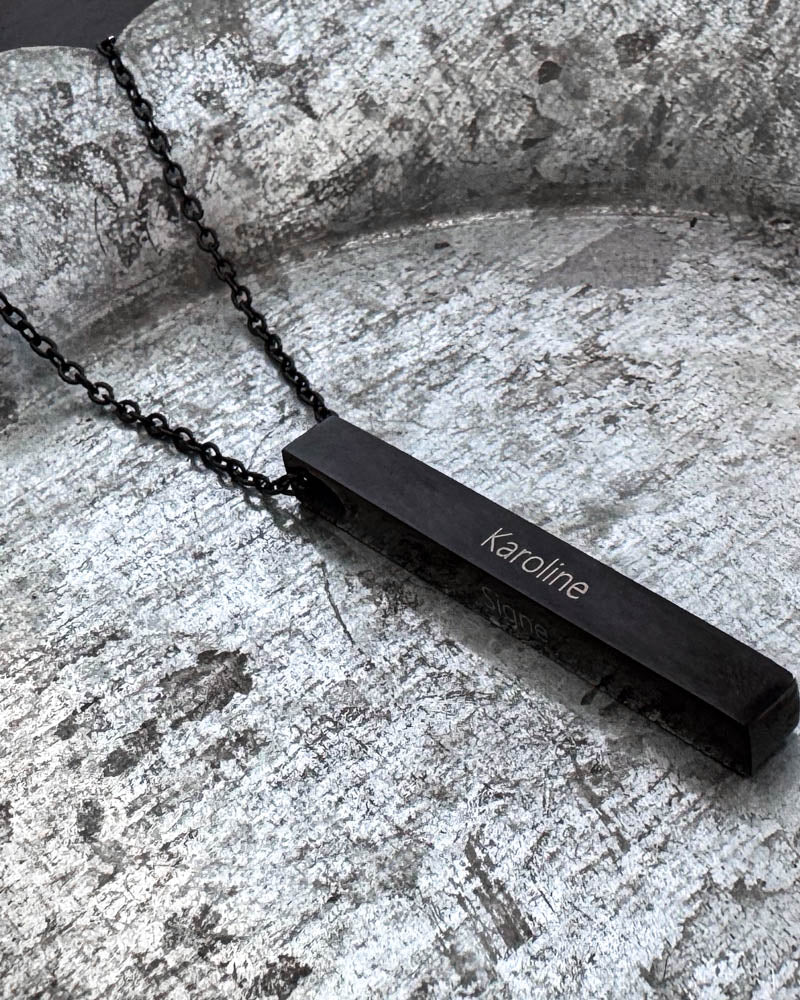 NEO name necklace in black stainless steel with the name "Karoline" engraved on an elongated bar, lying on a patinated zinc tray
