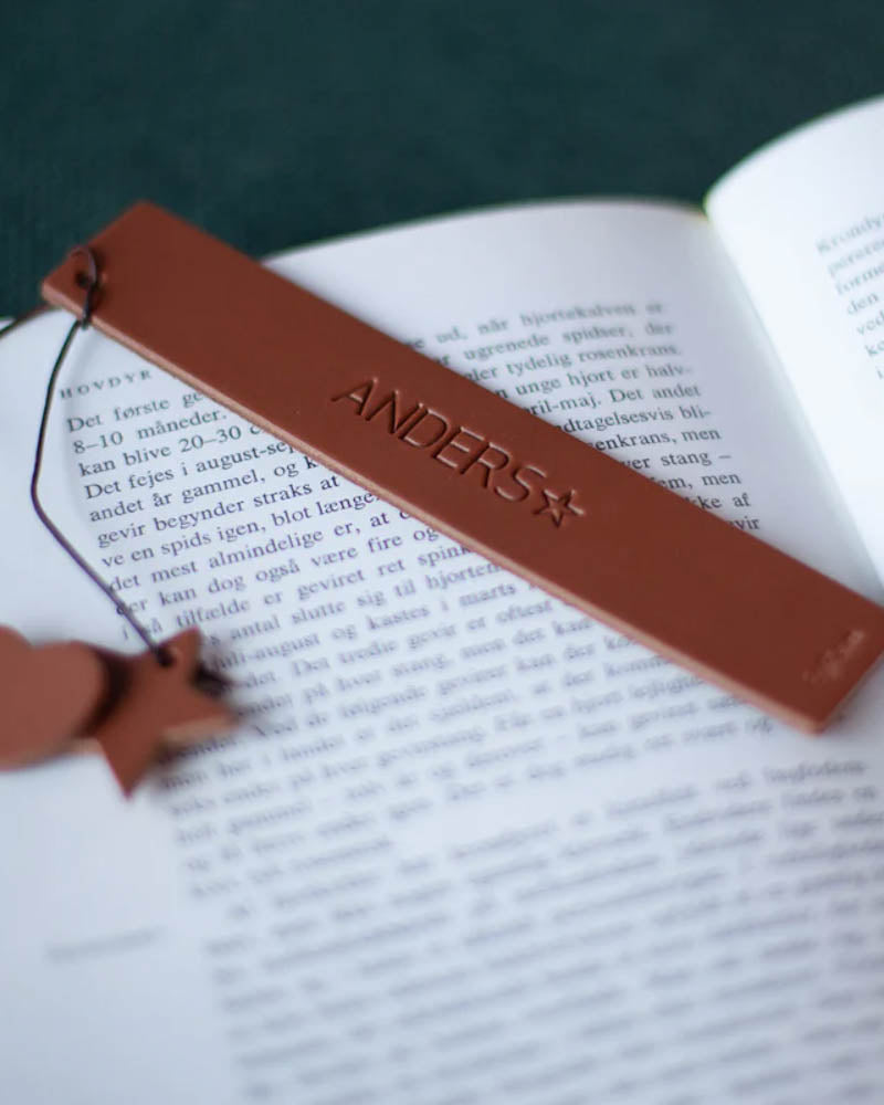PAGE bookmark in cognac leather with the name "Anders" and a star engraved, lying in an open book