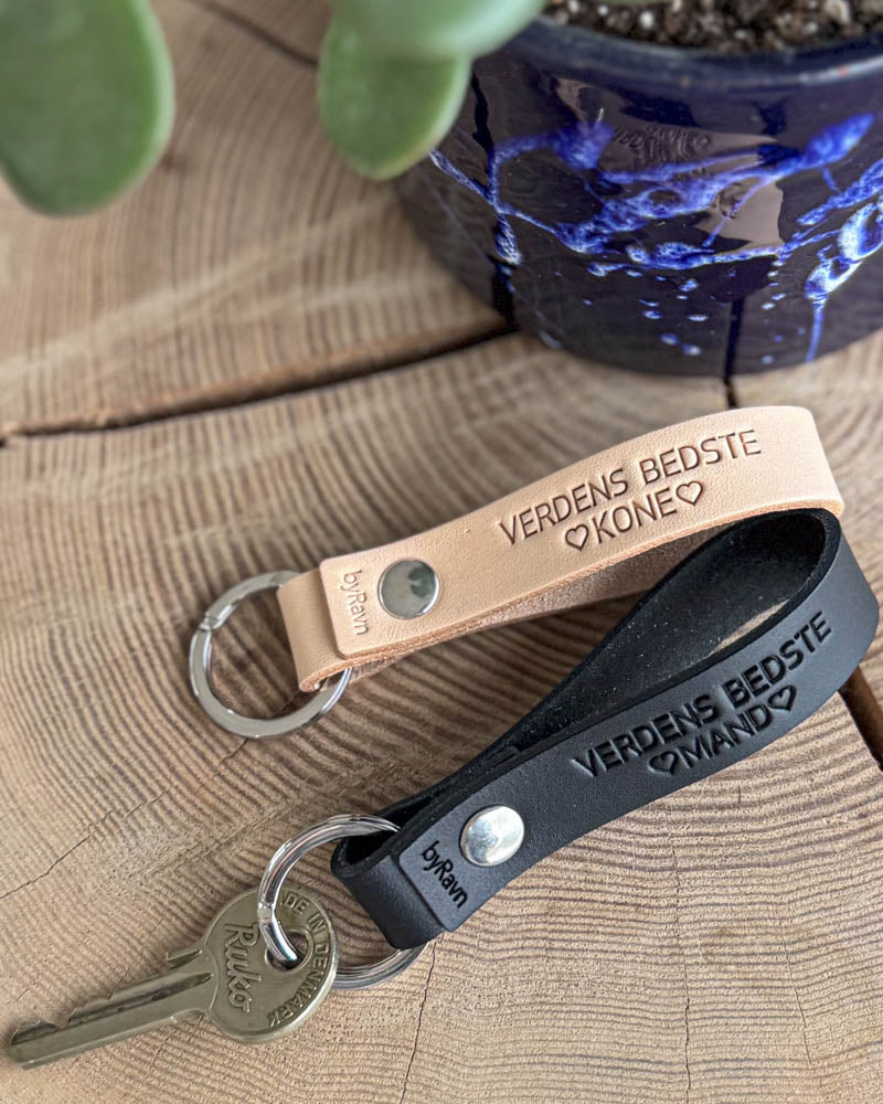 RAVN leather keyrings in black and natural engraved "Verdens bedste mand" and "Verdens bedste kone" ♡ on a wooden table by a blue pot
