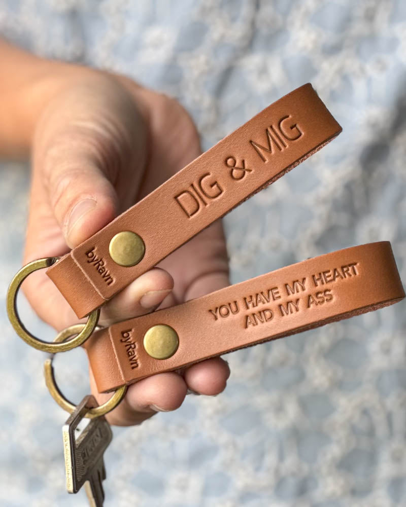 Two RAVN leather keyrings in cognac held in hand with the engravings "Dig & mig" and "You have my heart and my ass"