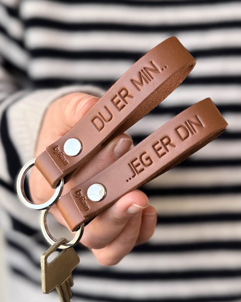 RAVN leather keyrings in cognac as a couple's set with the engravings "Du er min.." and "..jeg er din", held in hand with key