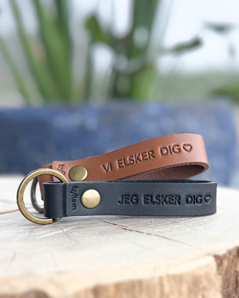 RAVN leather keyrings in cognac and black on a wooden slice with the engravings "Vi elsker dig" ♡ and "Jeg elsker dig" ♡