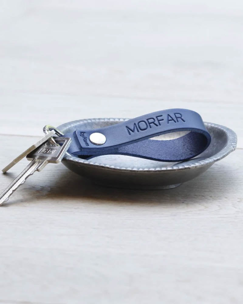 RAVN leather keyring in navy blue with the engraving "Morfar" and keys in a small metal bowl on a light wooden floor