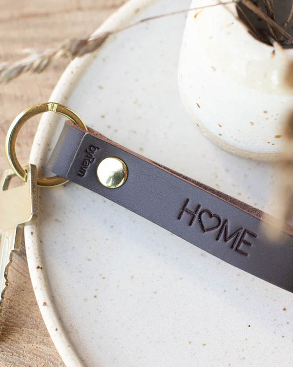 RAVN leather keyring in dark brown with golden brass rivet and the engraving "Home" with a heart, on a light ceramic dish with key