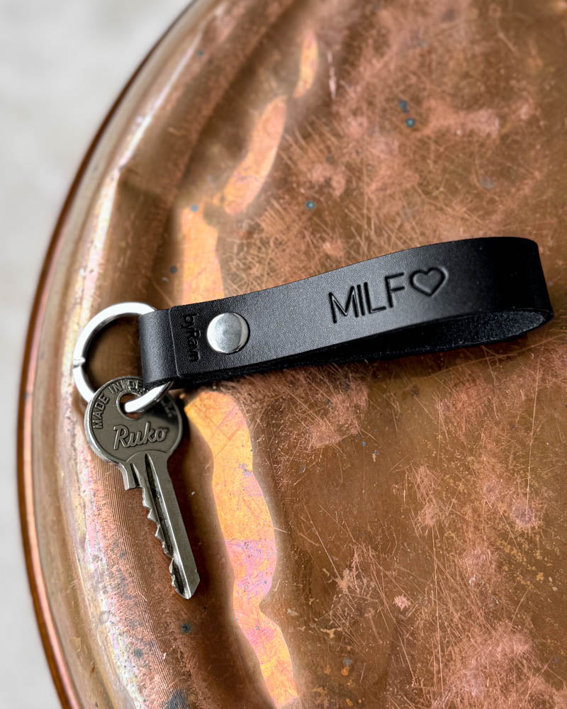 RAVN leather keyring in dark leather with the engraving "MILF" ♡ and key, lying on a patinated copper tray