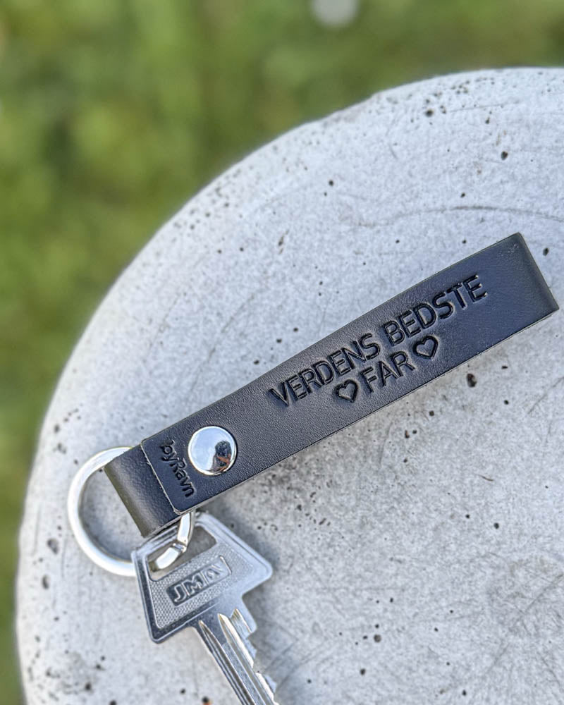 RAVN leather keyring in black with the engraving "Verdens bedste far" ♡ and key, lying on a concrete disc in the grass