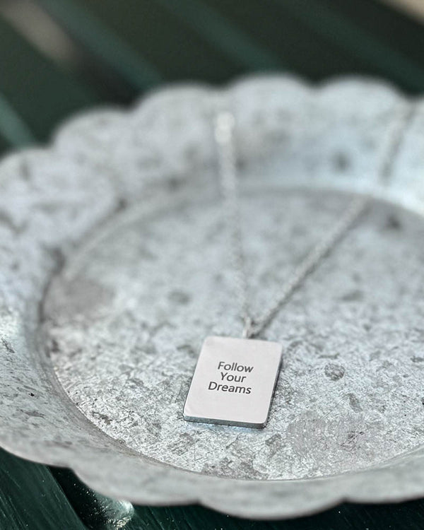 RAZ necklace in silver-coloured stainless steel with "Follow Your Dreams" engraved on rectangular plate, on white-patinated zinc tray