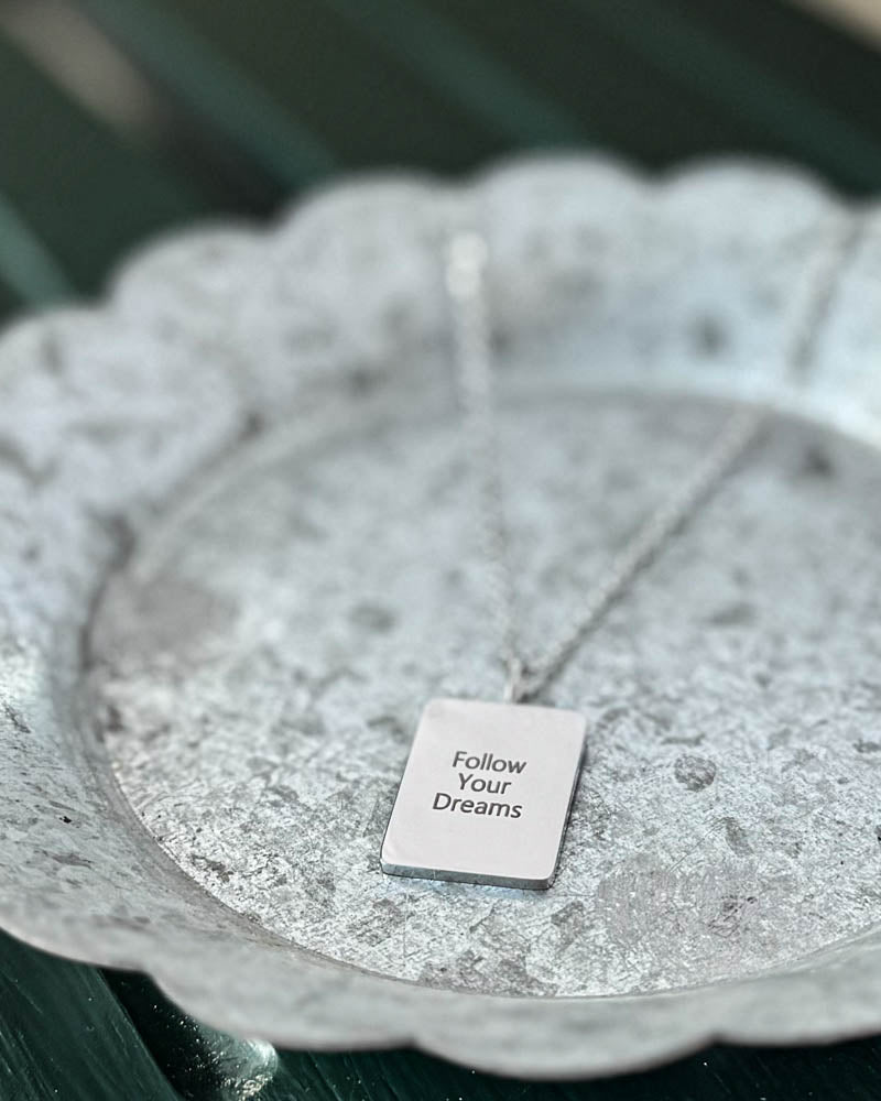 RAZ necklace in silver-coloured stainless steel with "Follow Your Dreams" engraved on rectangular plate, on white-patinated zinc tray