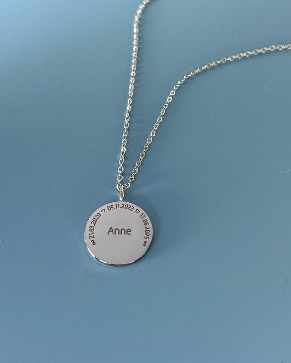 SIA CIRCLE name necklace, silver-coloured steel, "Anne" and dates "21.03.2020 ♡ 09.11.2022 ♡ 17.06.2025" in a circle, on blue backdrop