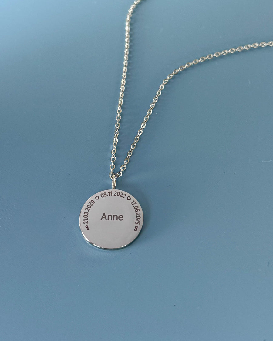 SIA CIRCLE name necklace, silver-coloured steel, "Anne" and dates "21.03.2020 ♡ 09.11.2022 ♡ 17.06.2025" in a circle, on blue backdrop