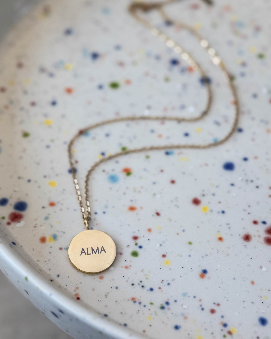 SIA NOTE name necklace in gold-plated stainless steel with the name "Alma" engraved on a round pendant, on a speckled ceramic plate