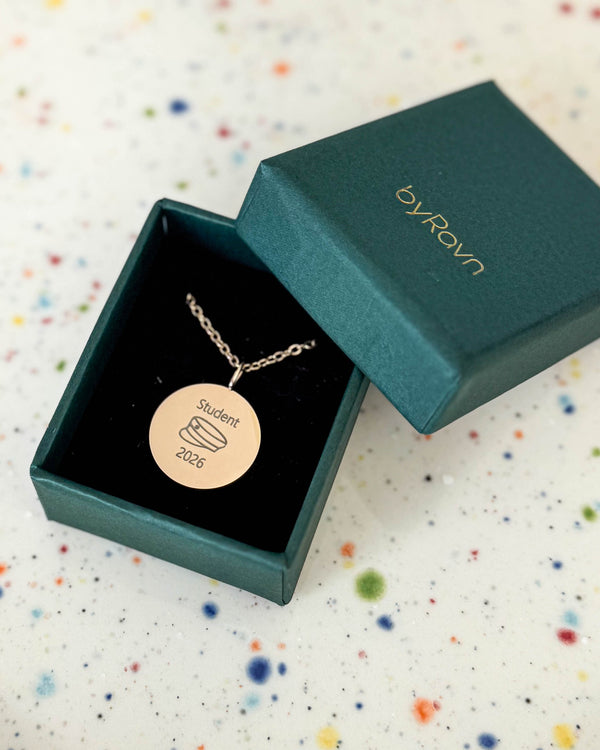 SIA MOMENTS necklace in gold-plated steel engraved with graduation cap and "Student 2026", in a green byRavn gift box on a speckled dish