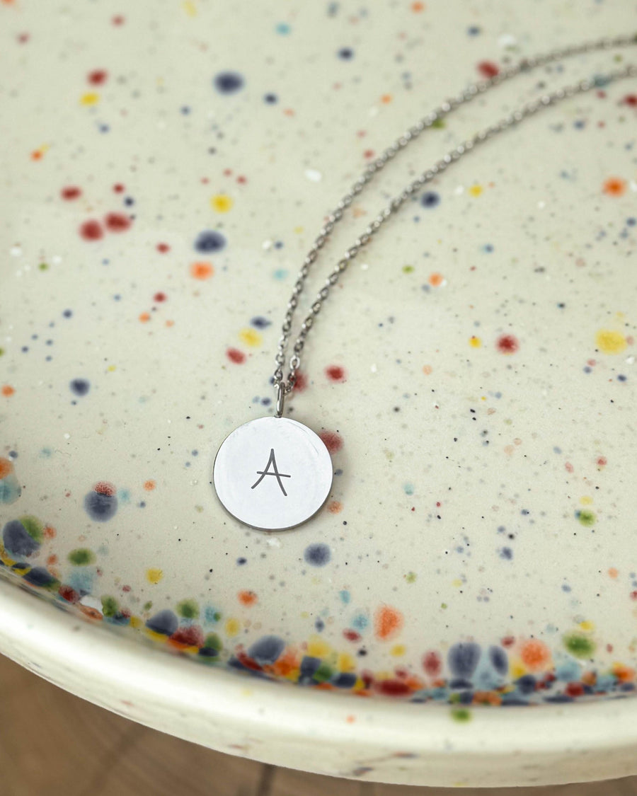 SIA LETTER SCRIPT necklace in silver-coloured stainless steel with handwritten letter "A" engraved, on a speckled ceramic dish