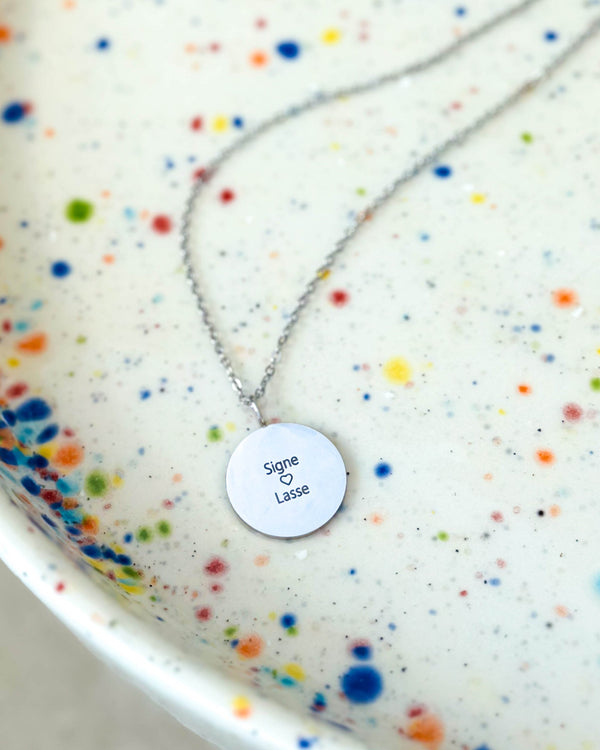 SIA QUOTE name necklace in silver-coloured stainless steel with the engraving "Signe ♡ Lasse" on a round pendant, on a speckled plate