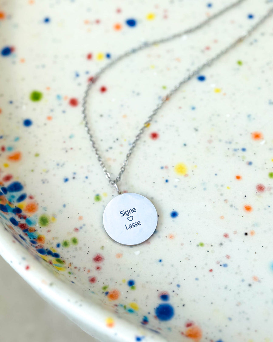 SIA QUOTE name necklace in silver-coloured stainless steel with the engraving "Signe ♡ Lasse" on a round pendant, on a speckled plate