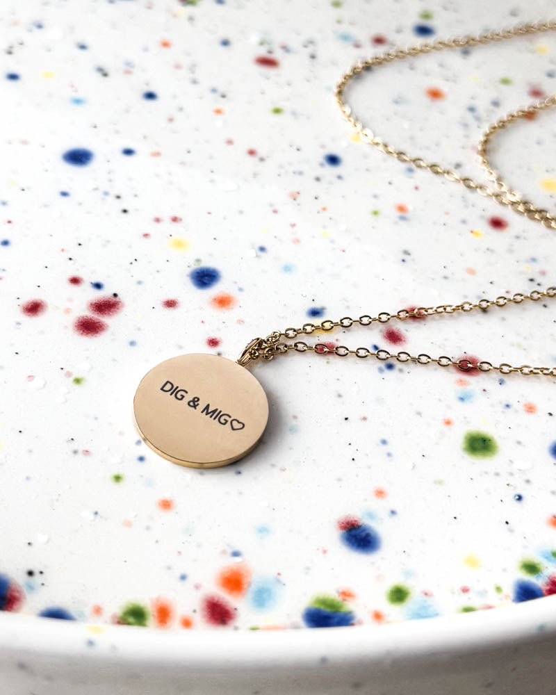 SIA QUOTE necklace in gold-plated stainless steel with the engraving "Dig & mig" ♡ on a round pendant, on a speckled ceramic plate