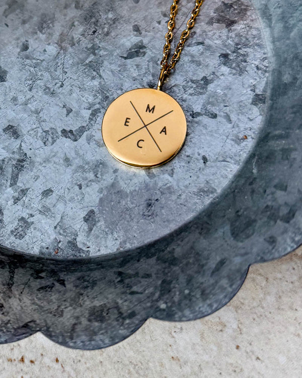 SIA X necklace in gold-plated stainless steel with the initials "M E A C" engraved in a cross on a round pendant, lying on a zinc tray
