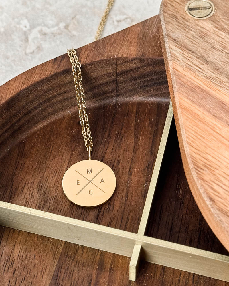 SIA X necklace in gold-plated stainless steel with the letters "M E A C" in a cross on a round pendant, in a wooden bowl with brass rim
