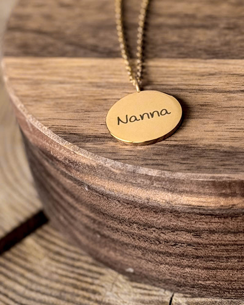 SIA NOTE SCRIPT name necklace in gold-plated steel with the name "Nanna" in handwriting engraved on a round pendant, on a round wooden dish