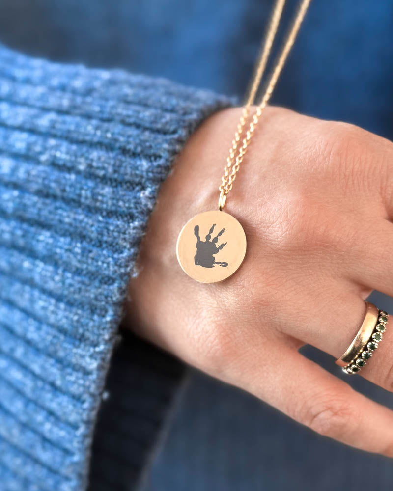 SIA MOTIF necklace in gold-plated stainless steel with a small handprint engraved on a round pendant, held in hand over blue knitwear
