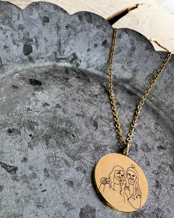 SIA PORTRAIT necklace in gold-plated stainless steel with line-drawn portrait of two women on a round pendant, lying on a zinc tray