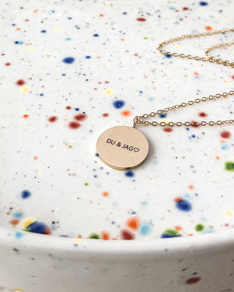 SIA QUOTE necklace in gold-plated stainless steel with the Swedish engraving "Du & jag" ♡ on a round pendant, on a speckled plate