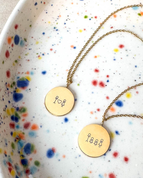 SIA STICK necklaces in gold-plated steel engraved with matchstick couple with heart and matchstick family, on a speckled ceramic dish