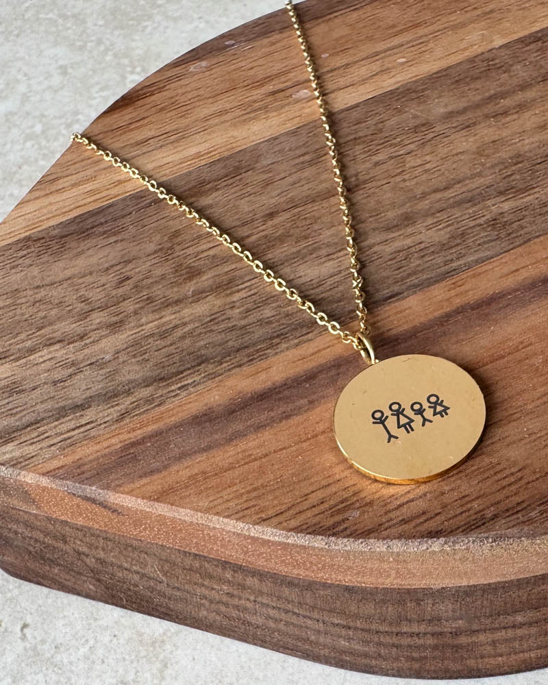 SIA STICK necklace in gold-plated stainless steel with a matchstick family of four engraved on a round pendant, on a dark wooden board