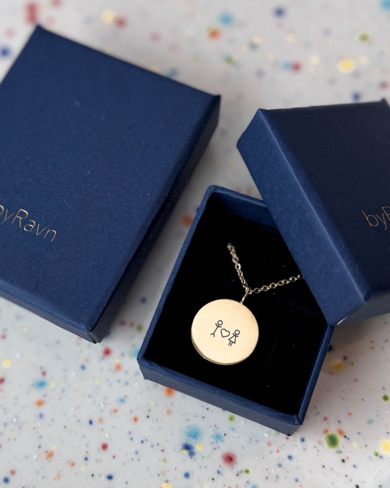 SIA STICK necklace in gold-plated steel engraved with matchstick couple and heart, in a navy blue byRavn gift box on a speckled dish