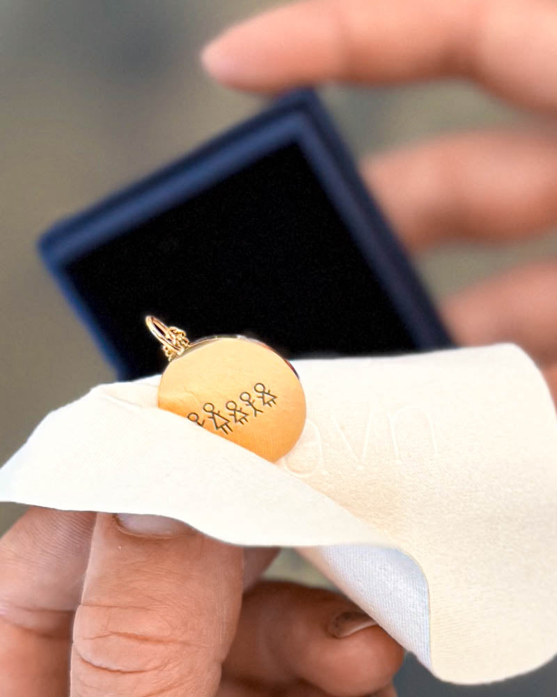 SIA STICK necklace in gold-plated stainless steel engraved with a matchstick family, held in hand on a polishing cloth by the gift box