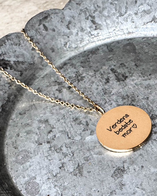SIA QUOTE SCRIPT necklace in gold-plated stainless steel engraved with the handwriting "Verdens bedste mor" ♡, lying on a zinc tray