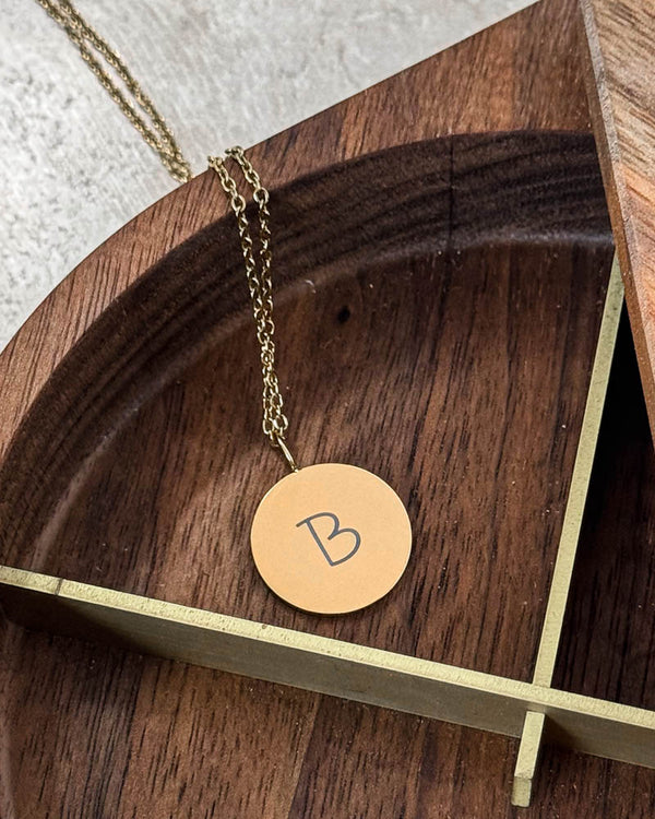 SIA LETTER SCRIPT necklace in gold-plated steel with handwritten letter "B" engraved on a round pendant, in a wooden bowl with brass rim