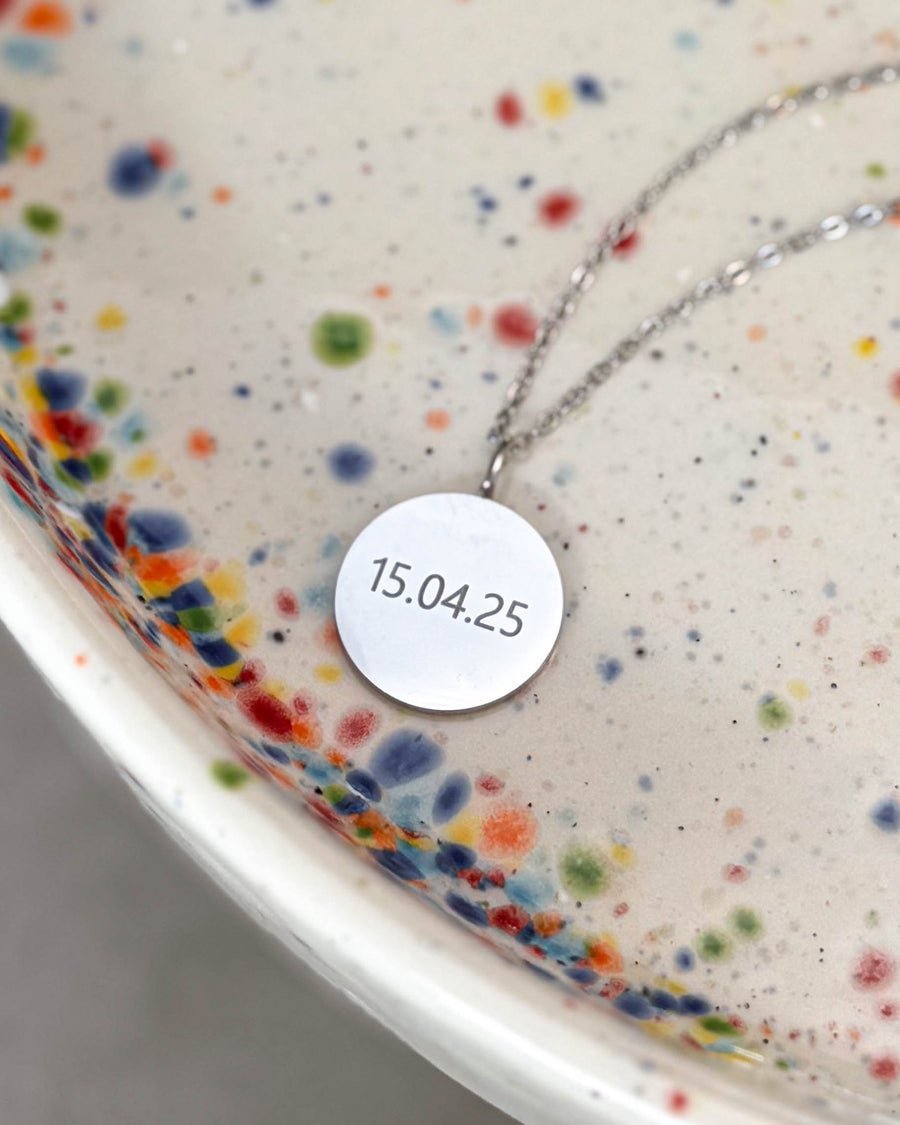 SIA NOTE necklace in silver-coloured stainless steel with the date "15.04.25" engraved on a round pendant, on a speckled ceramic dish