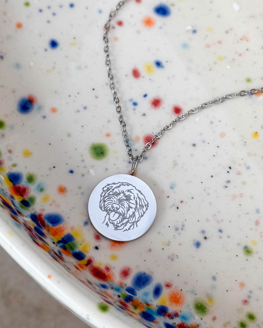 SIA PORTRAIT necklace in silver-coloured stainless steel with a drawing of a curly dog engraved on a round pendant, on a speckled dish