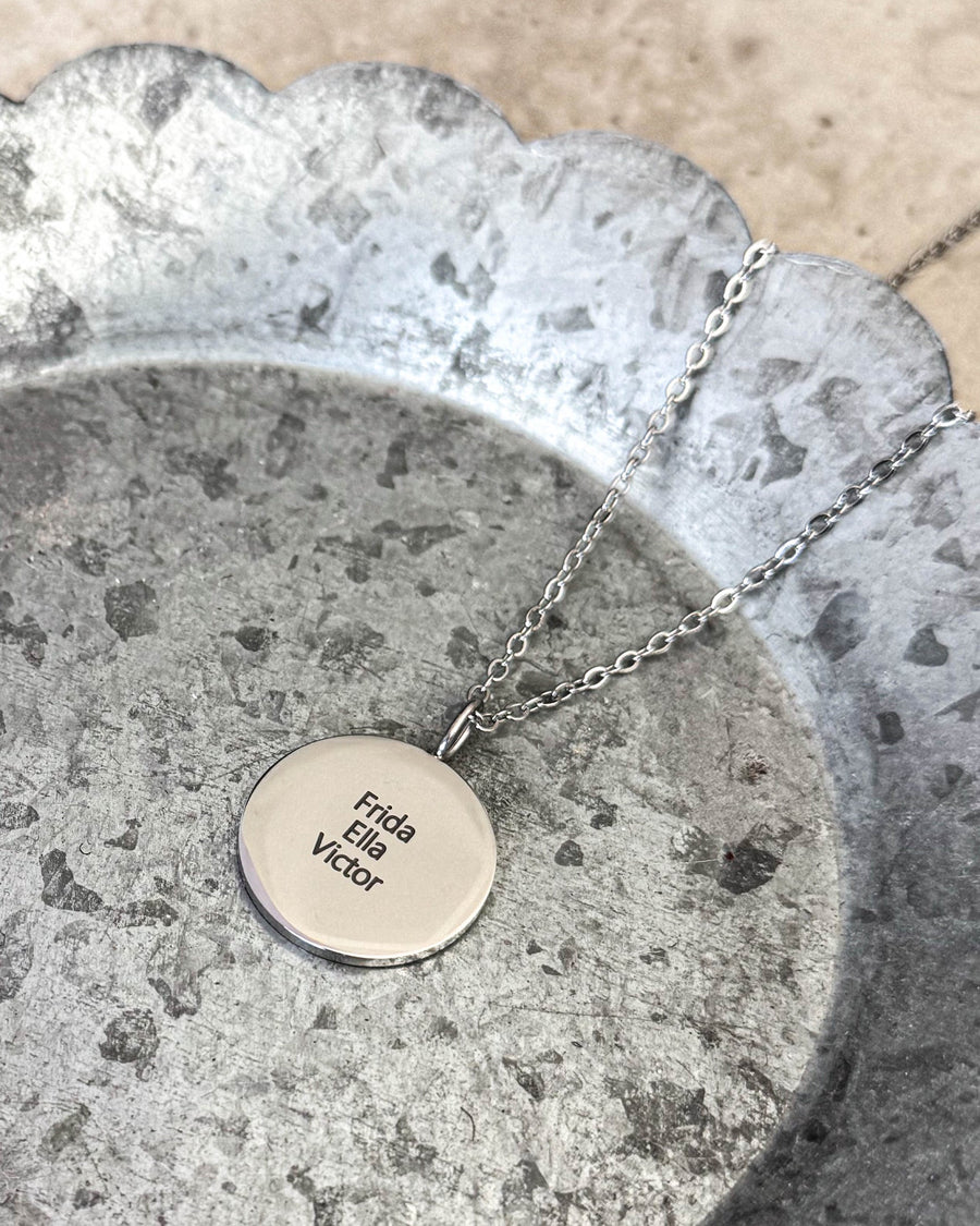 SIA QUOTE name necklace in silver-coloured stainless steel with the names "Frida, Ella, Victor" engraved, lying on a zinc tray