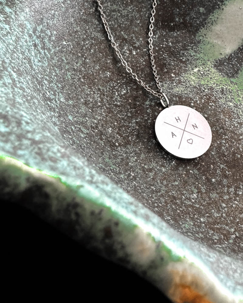 SIA X necklace in silver-coloured steel with the initials "H A N ♡" engraved in a cross on a round pendant, in a green ceramic bowl