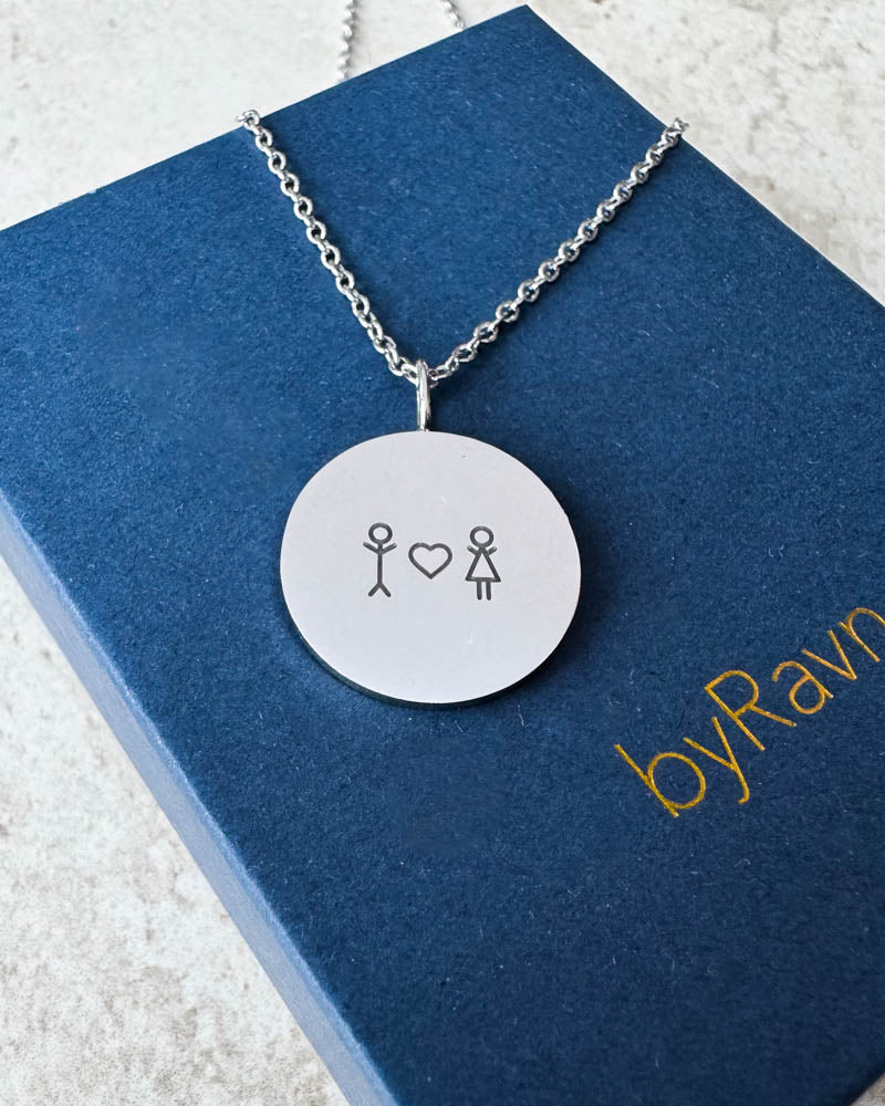 SIA STICK necklace in silver-coloured steel engraved with matchstick couple and heart, lying on a navy blue byRavn gift box
