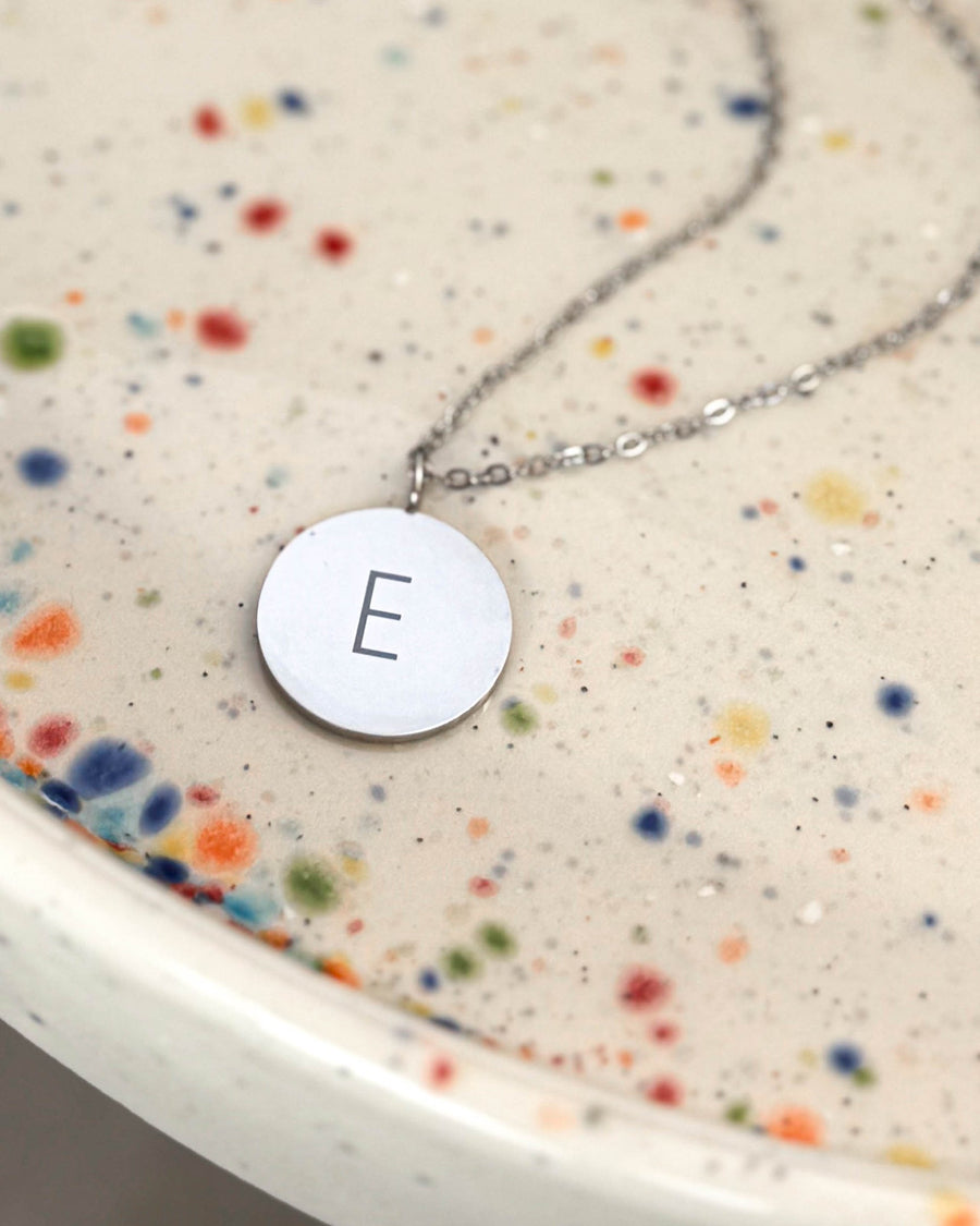 SIA LETTER necklace in silver-coloured stainless steel with the letter "E" engraved on a round pendant, on a speckled ceramic dish