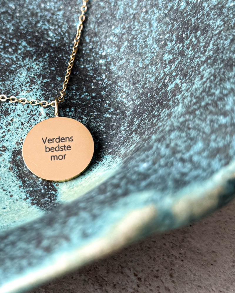 SIA QUOTE necklace in gold-plated stainless steel with the engraving "Verdens bedste mor" on a round pendant, in a turquoise ceramic bowl