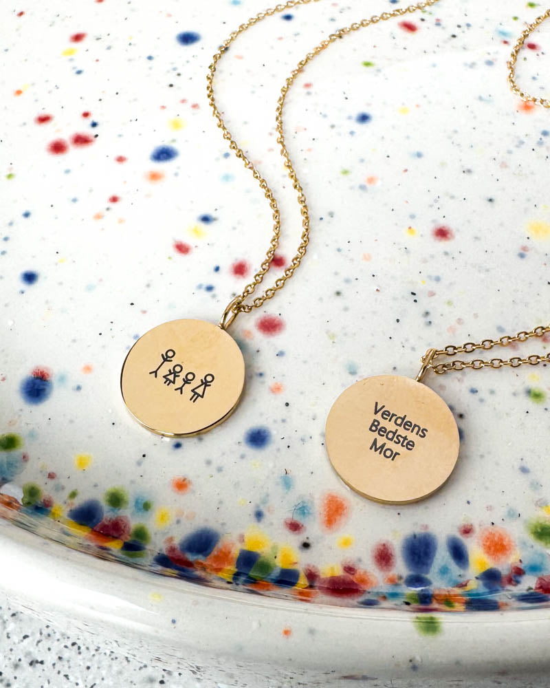 SIA STICK necklaces in gold-plated steel with matchstick family and the engraving "Verdens Bedste Mor", on a speckled ceramic dish