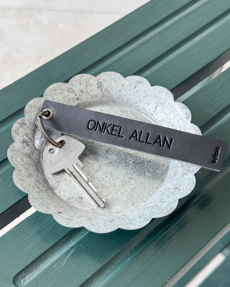 SOLO Large keyring in black leather with the engraving "Onkel Allan" and key in a zinc bowl on a green bench
