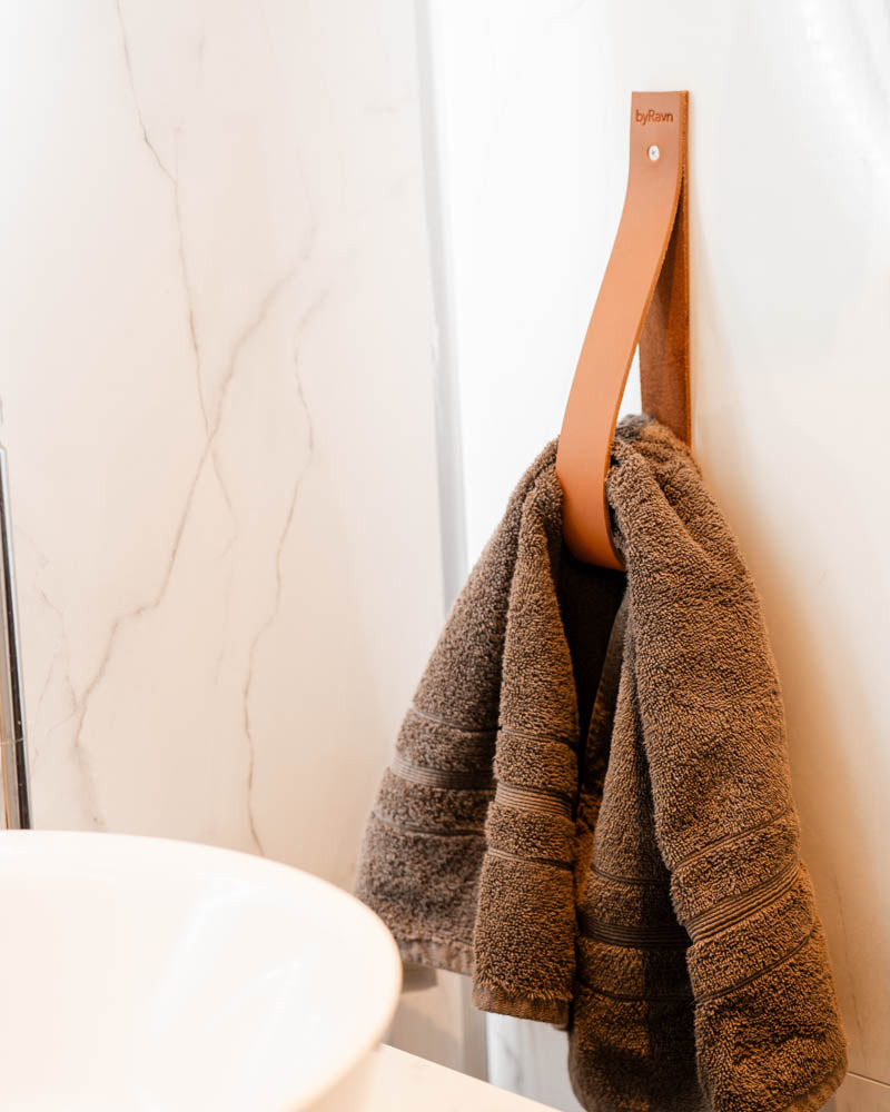 STACK magazine holder in cognac leather with byRavn embossing on a wall with a brown towel by a washbasin