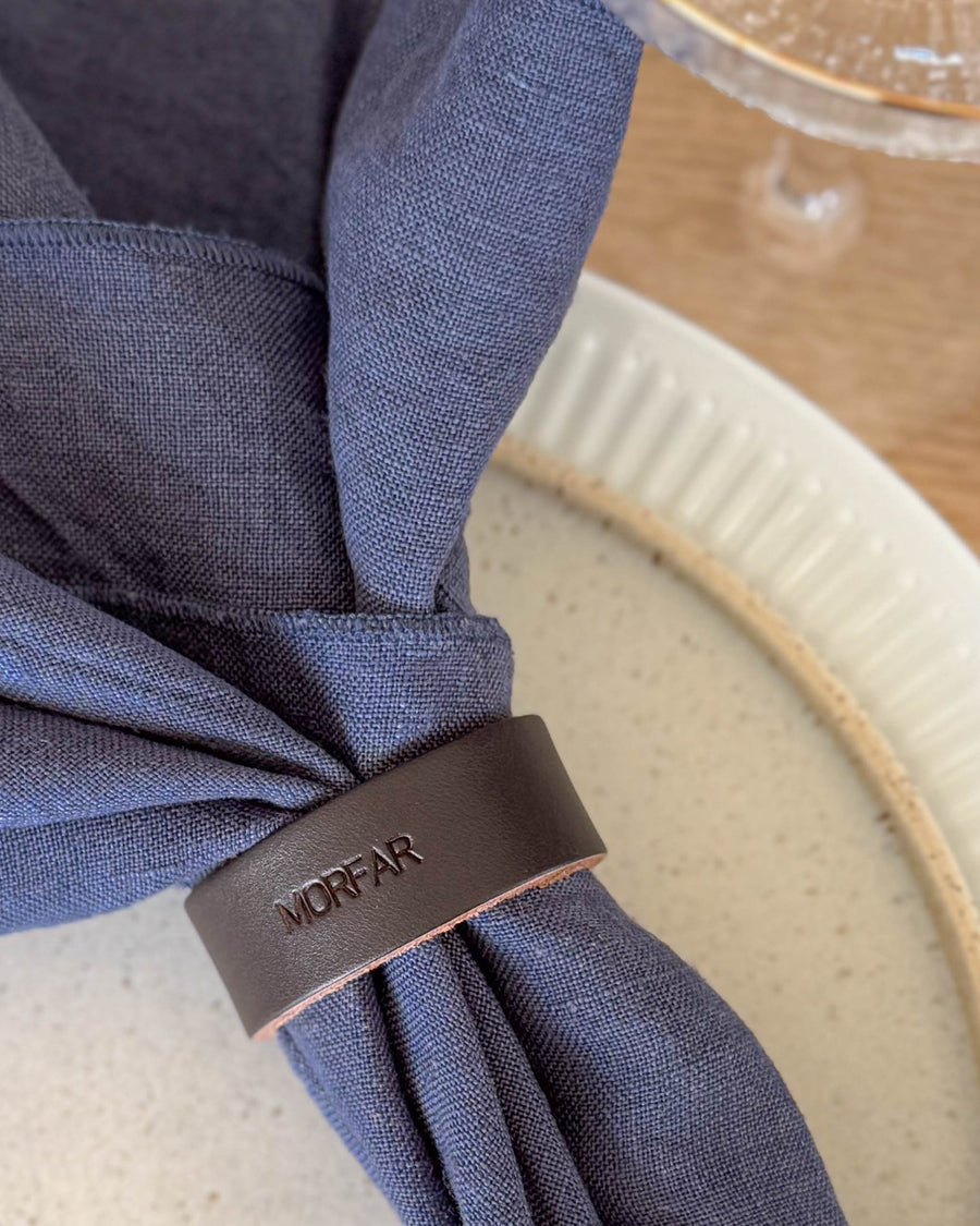 KNOT napkin ring place card in dark brown leather with the engraving "Morfar" around a navy linen napkin on a ribbed plate