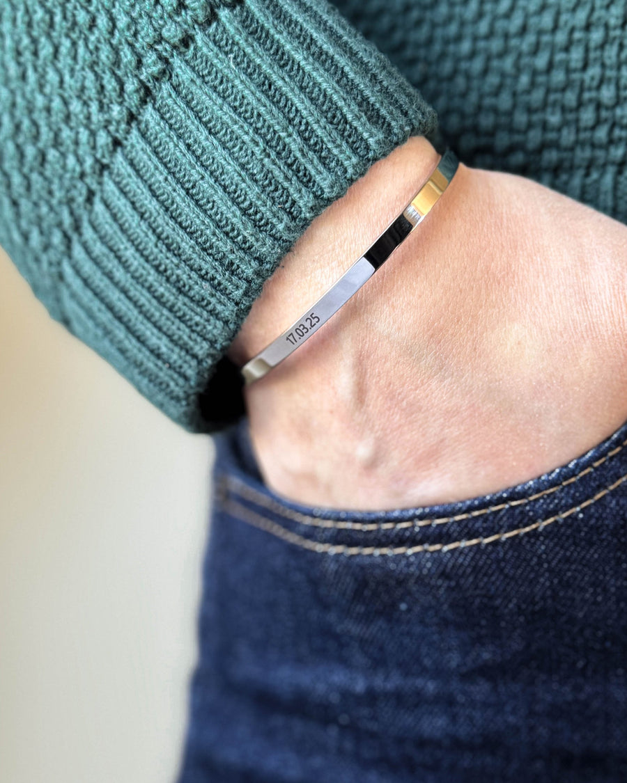 VENUS bracelet in silver-coloured stainless steel with the date "17.03.25" engraved, hand in dark jeans pocket, green knit